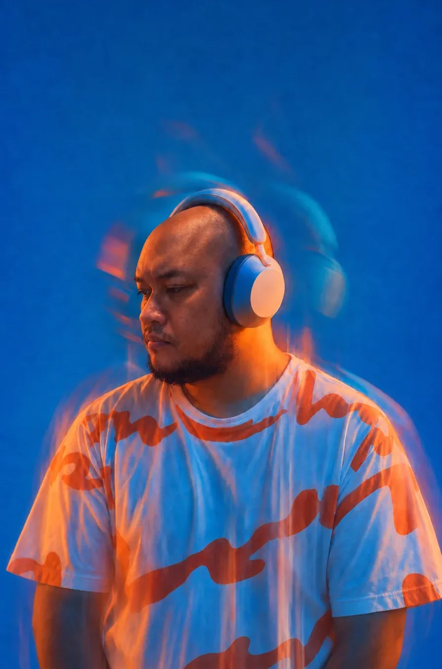 Arya Pradana in pale blue over-ear headphones and an orange-patterned t-shirt, lit against a deep cobalt backdrop with warm orange motion-blur streaks trailing around him.